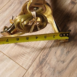 May include: A gold-colored decorative lamp base with a textured design. The lamp base is approximately 7 inches long. The electrical cord is coiled near the base. A yellow measuring tape is in the foreground.