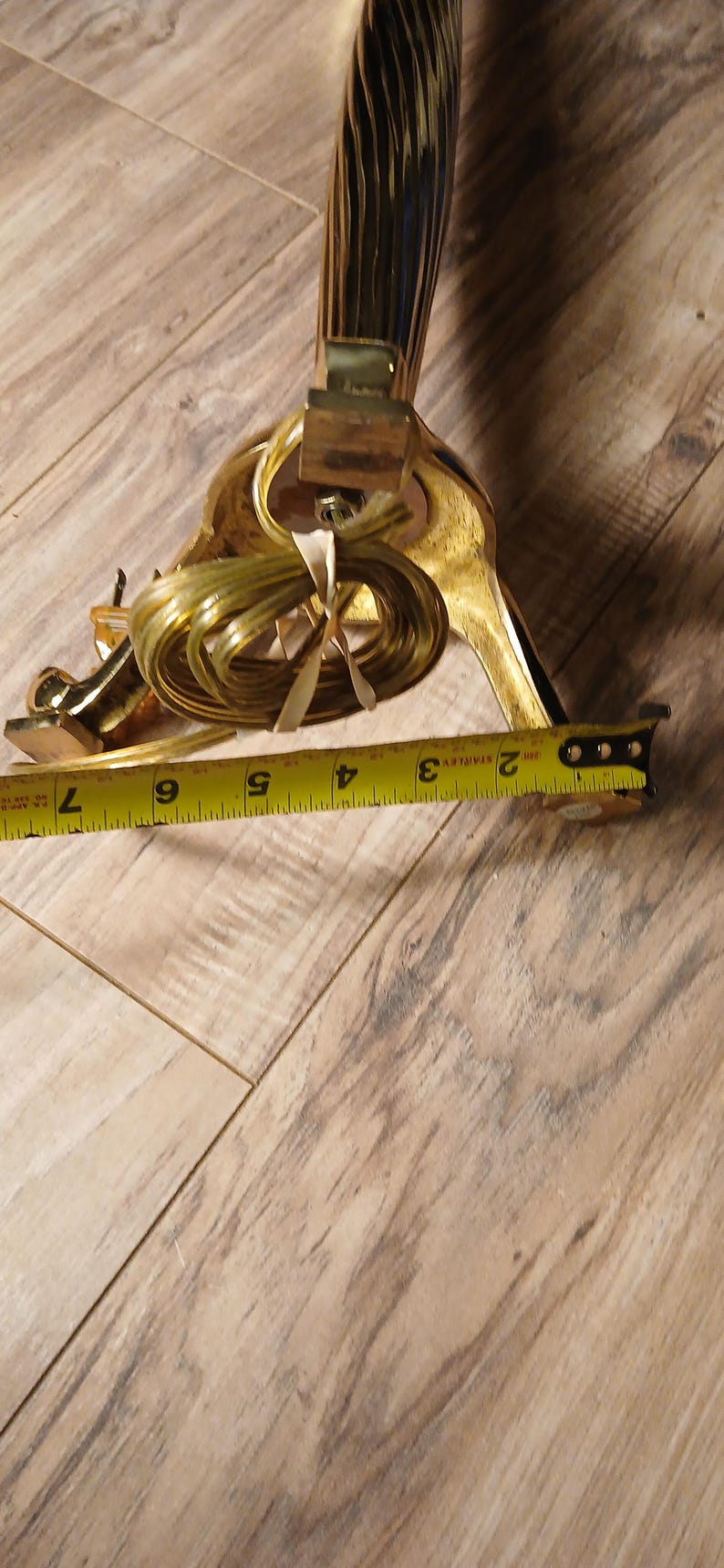 May include: A gold-colored decorative lamp base with a textured design. The lamp base is approximately 7 inches long. The electrical cord is coiled near the base. A yellow measuring tape is in the foreground.