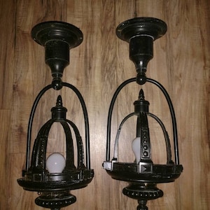 May include: Two black metal ceiling light fixtures with cage-like shades and white light bulbs. The fixtures have a decorative design with a pointed finial at the bottom.