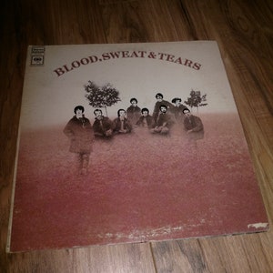 May include: A vintage vinyl record album cover featuring a group of men in a field with trees. The album cover is titled "Blood, Sweat & Tears".