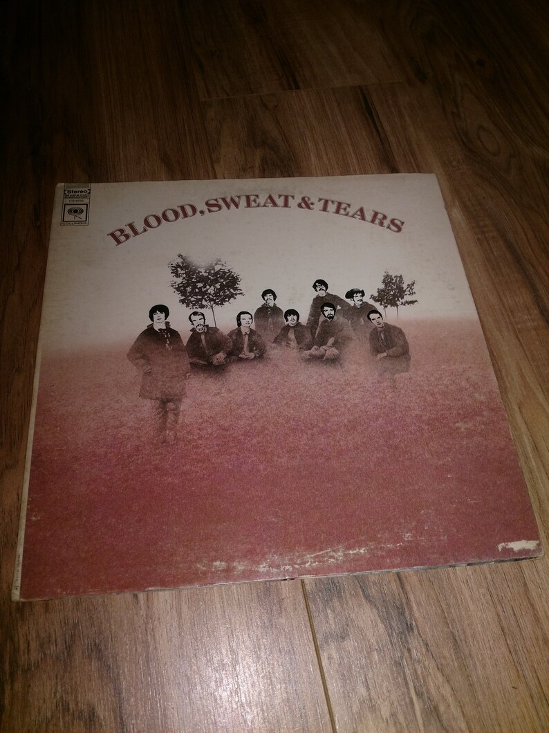 May include: A vintage vinyl record album cover featuring a group of men in a field with trees. The album cover is titled "Blood, Sweat & Tears".