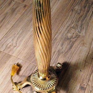 May include: A gold-colored electric candle lamp with a fluted column and a scalloped candle holder. The base has a decorative design and three feet. A cream-colored candle is in the holder.
