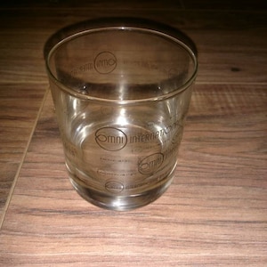 May include: A clear glass tumbler with the words "OMNI INTERNATIONAL" printed around the sides. The glass is cylindrical with a slightly wider base and sits on a wooden surface.