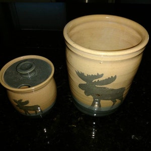 May include: Two ceramic jars with moose silhouettes. The smaller jar has a lid and is light brown with a blue lid. The larger jar is a light brown color with a blue moose silhouette.