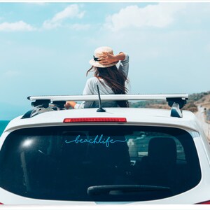 May include: White car with a roof rack and the word "beachlife" in a cursive font on the rear window. A person wearing a hat is visible, looking out at the ocean and sky. The scene evokes a sense of travel and leisure.
