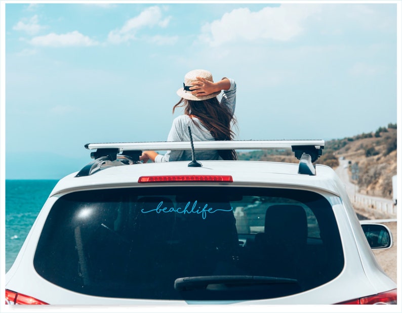 May include: White car with a roof rack and the word "beachlife" in a cursive font on the rear window. A person wearing a hat is visible, looking out at the ocean and sky. The scene evokes a sense of travel and leisure.