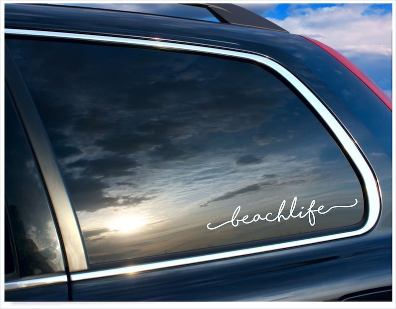 May include: A black car window with the white cursive text "beachlife" applied. The window reflects a cloudy sky with a setting sun, creating a serene and beachy vibe.