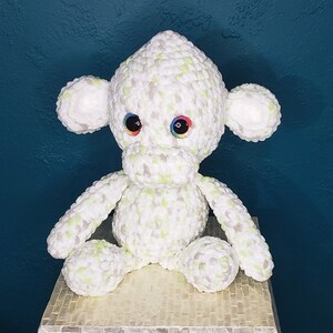 May include: A white crocheted monkey toy with green accents and colorful eyes. The monkey is sitting on a light brown surface.