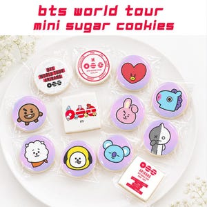May include: A white plate holds a collection of mini sugar cookies, each individually wrapped. The cookies feature colorful designs and text, including "BTS WORLD TOUR" and various character illustrations. The cookies are round and square.
