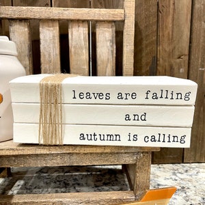 May include: A stack of three white books with the text "leaves are falling and autumn is calling" printed on them. The books are tied together with twine.