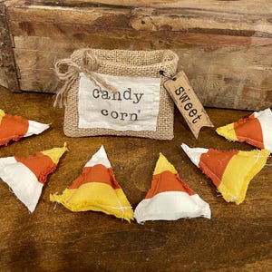 May include: A rustic burlap bag labeled "candy corn" with a small tag that says "sweet". Several fabric candy corn pieces in yellow, orange, and white are scattered around the bag. The background includes a wooden box.