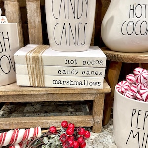 May include: A stack of three white books with black text that reads "hot cocoa", "candy canes", and "marshmallows". The books are stacked on a wooden shelf with a white mug that says "candy canes" and a white pitcher that says "hot cocoa" on the shelf. There are red and white candy canes and red berries in the foreground.