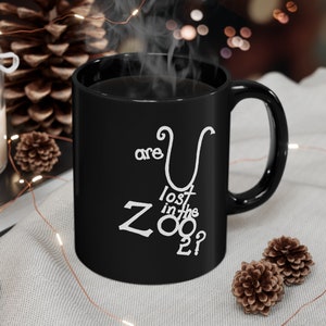 May include: Black coffee mug with white text that reads "are you lost in the zoo?" The mug has a white handle and is sitting on a white surface.