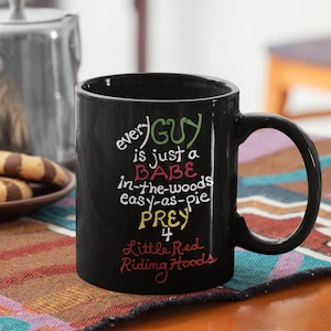 Peut inclure: Mug à café noir avec un dicton humoristique en texte coloré : « every GUY is just a BABE in-the-woods easy-as-pie PREY 4 Little Red Riding Hoods »