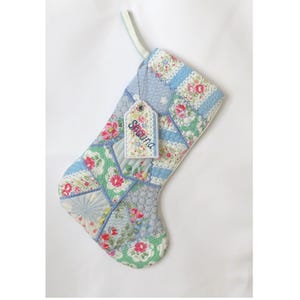 May include: A quilted Christmas stocking featuring a patchwork design of light blue, white, and green floral patterns with various textures. A small, rectangular fabric tag is attached to the top, embroidered with the name Shauna in blue.