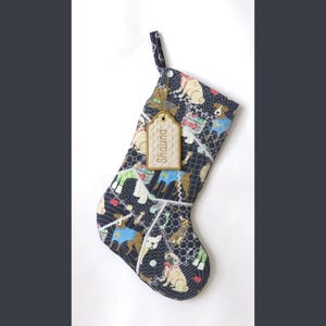 May include: A quilted Christmas stocking featuring a dark blue fabric with a repeating pattern of various dog breeds wearing sweaters and accessories. A beige rectangular tag with the name Shauna embroidered in brown hangs from the top loop.
