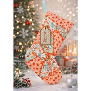 May include: A quilted Christmas stocking featuring a patchwork design of peach, orange, and teal fabric with star and floral patterns. A light blue ribbon loop holds a rectangular beige tag with the embroidered name Shauna in brown thread.