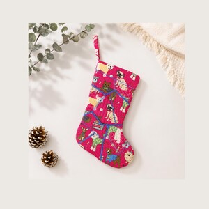 May include: A quilted Christmas stocking in a vibrant pink fabric featuring a repeating pattern of various dog breeds, including pugs and terriers, wearing colourful sweaters and scarves. The stocking has a matching pink fabric hanging loop.