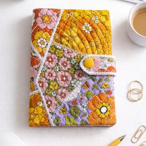 Puede incluir: Una funda de cuaderno floral acolchada en tonos naranja, amarillo, rosa y morado. La funda tiene un cierre de botón y costuras blancas. También se ven un bolígrafo amarillo, anillos dorados y clips.