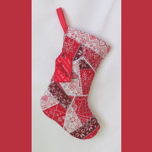 May include: A Christmas stocking featuring a patchwork design of red and white fabric with various geometric, snowflake, and heart patterns. A red fabric tag is attached to the top, embroidered with the name Shauna and the text Joy Noel.