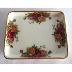 May include: A rectangular porcelain trinket tray with a cream-coloured base and a gold-trimmed edge. The tray features floral clusters of red, pink, and yellow roses with green leaves arranged along the four sides of the interior surface.
