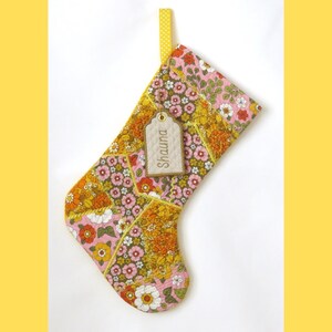 May include: A quilted Christmas stocking featuring a patchwork of floral fabric in shades of pink, orange, yellow, and green. A yellow fabric loop is attached to the top, and a beige rectangular tag with the name Shauna is tied to the stocking.