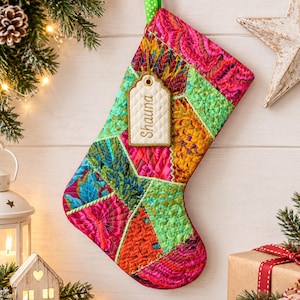 May include: A quilted Christmas stocking featuring a vibrant patchwork design of bright pink, orange, teal, and lime green fabric segments. A cream-coloured rectangular gift tag with a quilted texture hangs from the top, displaying the name Shauna.
