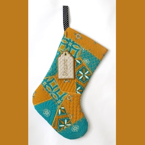 May include: A quilted Christmas stocking featuring a patchwork pattern of mustard yellow and teal blue fabric with geometric floral motifs. A black and white polka dot fabric loop is attached at the top, and a beige tag with the name Shauna is tied on.