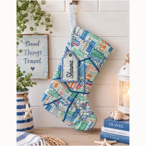 May include: A quilted Christmas stocking featuring a collage of vintage airmail postage stamps in shades of blue, green, and red. A white and black polka dot ribbon loop hangs the stocking, which includes a rectangular tag labelled Shauna.