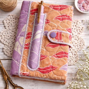 May include: Quilted journal with a floral pattern in shades of orange, red, and purple. The journal has a pen holder and a button closure. A gold pen is inserted in the pen holder. The journal is on a white wooden surface.