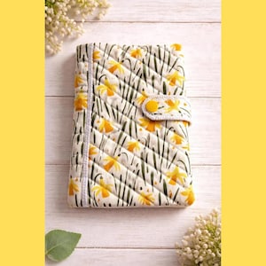 May include: A rectangular quilted fabric notebook cover featuring a repeating pattern of yellow daffodils and green stems on a white background. The cover includes a flap closure secured with a single yellow snap button and diagonal stitching.