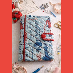 May include: A light blue quilted fabric cover featuring a vintage sewing pattern print in dark blue and red. The cover has a red fabric trim, a red button closure, and diagonal stitching. It is surrounded by sewing tools like needles and thread.