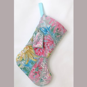 May include: A quilted Christmas stocking featuring a vibrant floral pattern in shades of pink, yellow, blue, and green. The stocking includes a light blue hanging loop and a rectangular fabric tag embroidered with the name Shauna in dark thread.