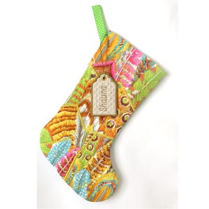 May include: A quilted Christmas stocking featuring a vibrant, multi-coloured feather pattern in shades of yellow, orange, pink, and blue. A green polka-dot fabric loop is attached to the top, and a beige quilted tag with the name Shauna is tied on.