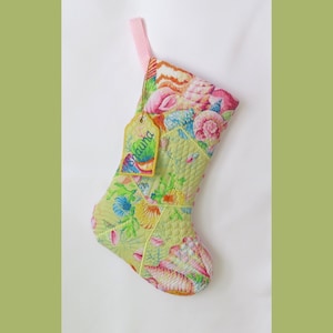 May include: A quilted Christmas stocking featuring a vibrant pattern of seashells and coral in shades of pink, yellow, and blue on a light green background. A small, matching fabric tag attached with a string displays the name Shauna in green.