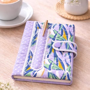 Könnte beinhalten: Ein gestepptes Notizbuch mit einem Blumenmuster in Blau, Grün und Gelb und einem lavendelfarbenen Tupfenmuster. Das Notizbuch hat einen Stift in der Wirbelsäule und einen Knopfverschluss. Eine Tasse Kaffee und Blumen sind im Hintergrund.