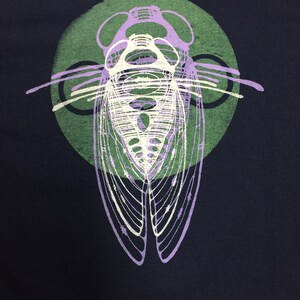 Pode incluir: Uma camiseta azul escura com uma estampa gr&aacute;fica de um inseto cigarra em um c&iacute;rculo. A cigarra &eacute; contornada em roxo e branco, e o c&iacute;rculo &eacute; verde.