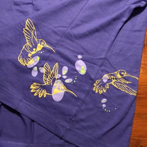 K&ouml;nnte beinhalten: Ein lila T-Shirt mit einem gelben Kolibri-Design. Der Kolibri fliegt durch ein Feld aus lila und gr&uuml;nen Punkten.