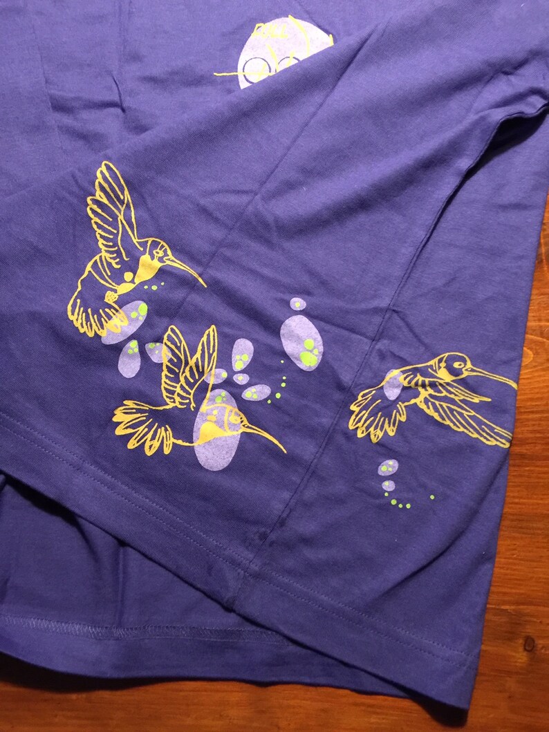 K&ouml;nnte beinhalten: Ein lila T-Shirt mit einem gelben Kolibri-Design. Der Kolibri fliegt durch ein Feld aus lila und gr&uuml;nen Punkten.