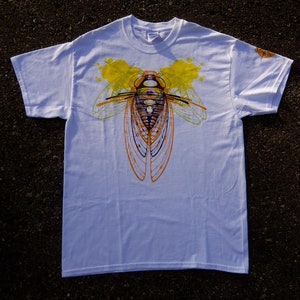 Pode incluir: Camiseta branca com uma estampa gr&aacute;fica de uma cigarra em amarelo, laranja e preto. A cigarra &eacute; contornada em preto e tem um corpo roxo.