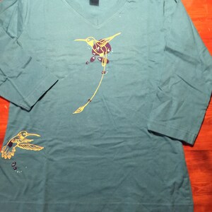 K&ouml;nnte beinhalten: Tealblaues Langarm-T-Shirt mit einem gelben Kolibri-Motiv. Der Kolibri hat einen langen Schwanz und ist von kleinen violetten Punkten umgeben.
