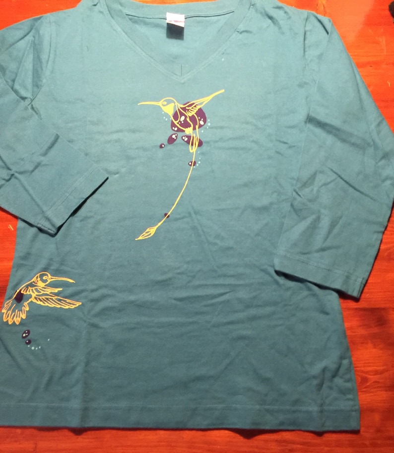 K&ouml;nnte beinhalten: Tealblaues Langarm-T-Shirt mit einem gelben Kolibri-Motiv. Der Kolibri hat einen langen Schwanz und ist von kleinen violetten Punkten umgeben.