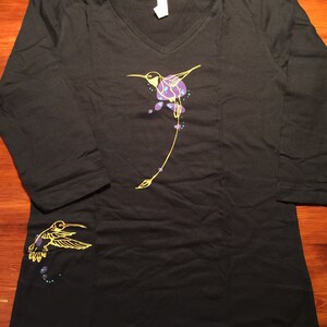 K&ouml;nnte beinhalten: Ein schwarzes Langarm-T-Shirt mit V-Ausschnitt und einem gelben Kolibri-Design. Der Kolibri ist in Gelb umrandet und hat lila und blaue Akzente. Auf dem linken &Auml;rmel befindet sich ein kleineres Kolibri-Design.