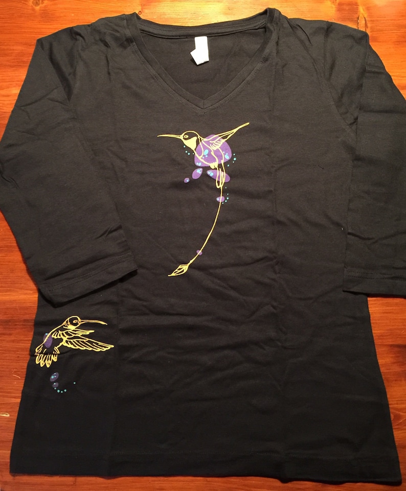 K&ouml;nnte beinhalten: Ein schwarzes Langarm-T-Shirt mit V-Ausschnitt und einem gelben Kolibri-Design. Der Kolibri ist in Gelb umrandet und hat lila und blaue Akzente. Auf dem linken &Auml;rmel befindet sich ein kleineres Kolibri-Design.