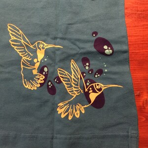 K&ouml;nnte beinhalten: Ein blaues T-Shirt mit einem goldenen Kolibri-Motiv. Der Kolibri fliegt mit ausgebreiteten Fl&uuml;geln. Um den Kolibri herum befinden sich violette und gr&uuml;ne Flecken.