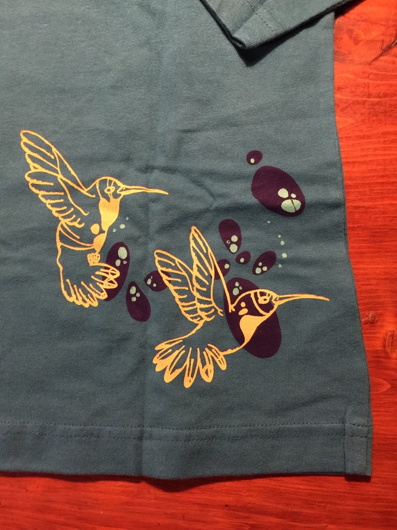 K&ouml;nnte beinhalten: Ein blaues T-Shirt mit einem goldenen Kolibri-Motiv. Der Kolibri fliegt mit ausgebreiteten Fl&uuml;geln. Um den Kolibri herum befinden sich violette und gr&uuml;ne Flecken.