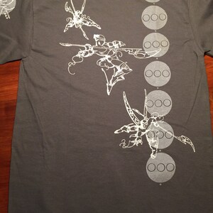 Op de afbeelding: Een donkergrijs T-shirt met een wit grafisch ontwerp van drie gestileerde figuren met vleugels en een reeks witte cirkels aan de rechterkant van het shirt.