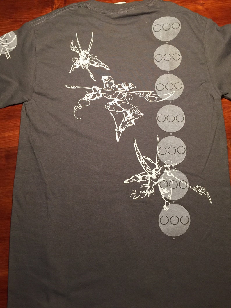 Op de afbeelding: Een donkergrijs T-shirt met een wit grafisch ontwerp van drie gestileerde figuren met vleugels en een reeks witte cirkels aan de rechterkant van het shirt.