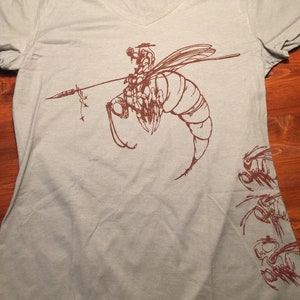 May include: A light gray t-shirt with a brown graphic of a bee with a rider on its back. The bee is flying and has a spear in its front legs. There are smaller bee graphics on the right side of the shirt.