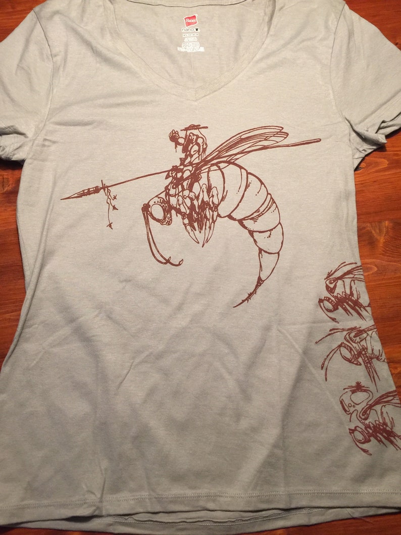 May include: A light gray t-shirt with a brown graphic of a bee with a rider on its back. The bee is flying and has a spear in its front legs. There are smaller bee graphics on the right side of the shirt.